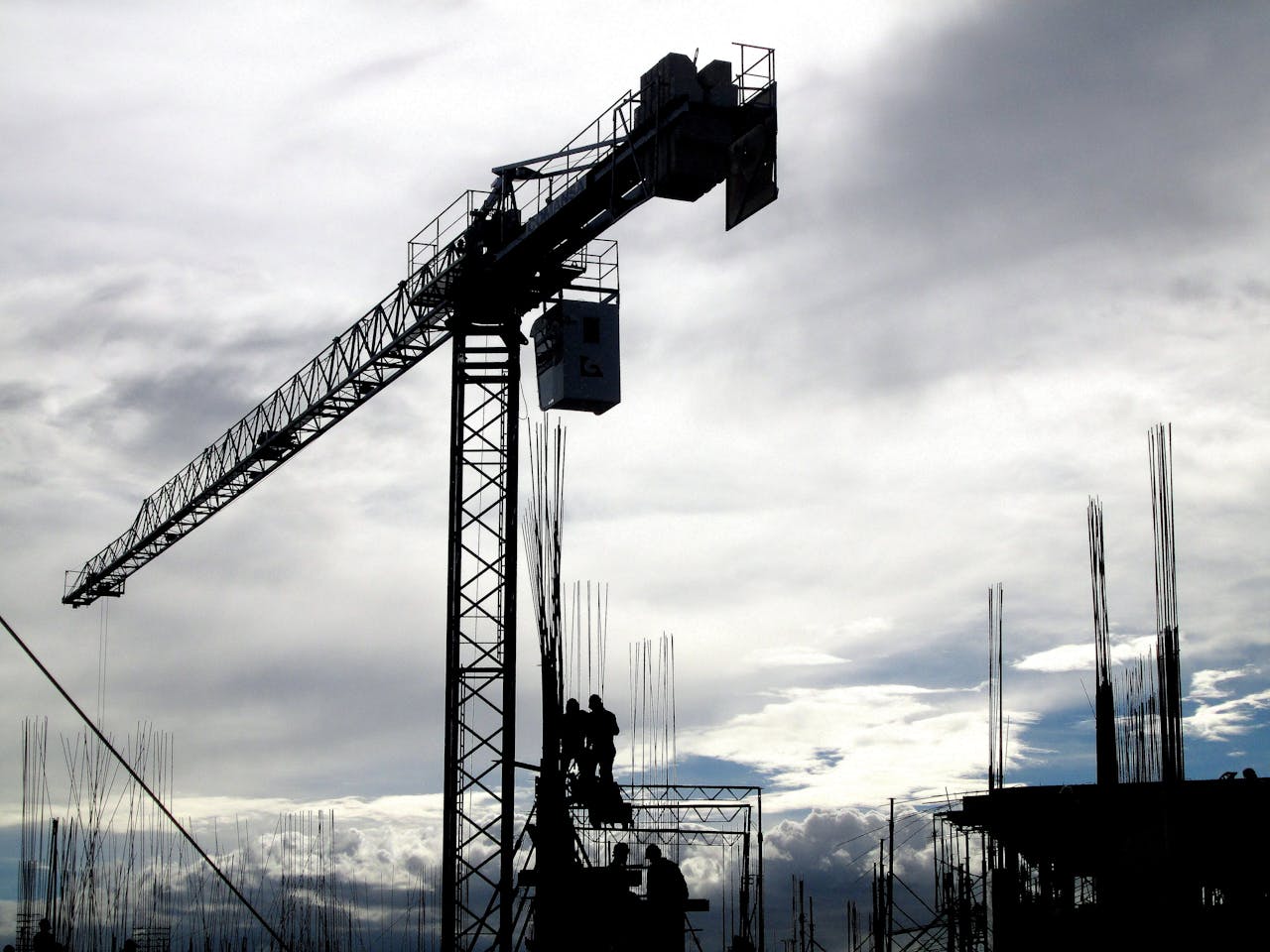 Silhouetted construction crane and workers against a cloudy sky during the day.