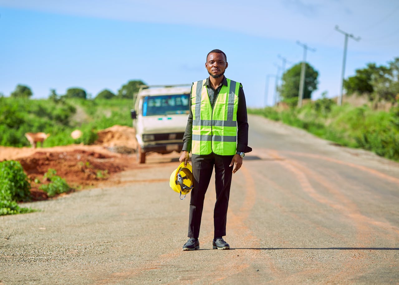 A construction worker in a safety vest stands on a rural road in Lafia, Nigeria, holding a helmet.