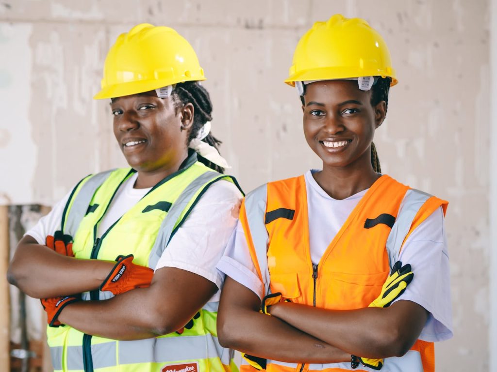 Two female construction workers wearing protective gear and smiling confidently indoors.